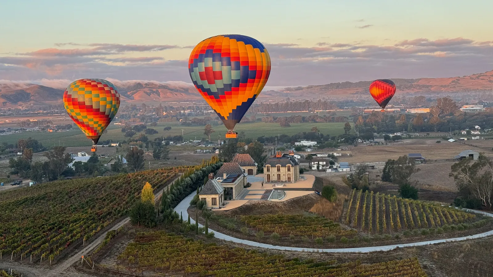 Three hot air balloons floating over Sonoma vineyard rows and a winery estate at sunrise in late summer